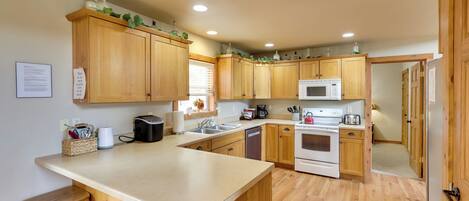 A kitchen with wooden cabinets, a microwave, a stove, and a sink. There are stools at the counter and a basket with utensils.