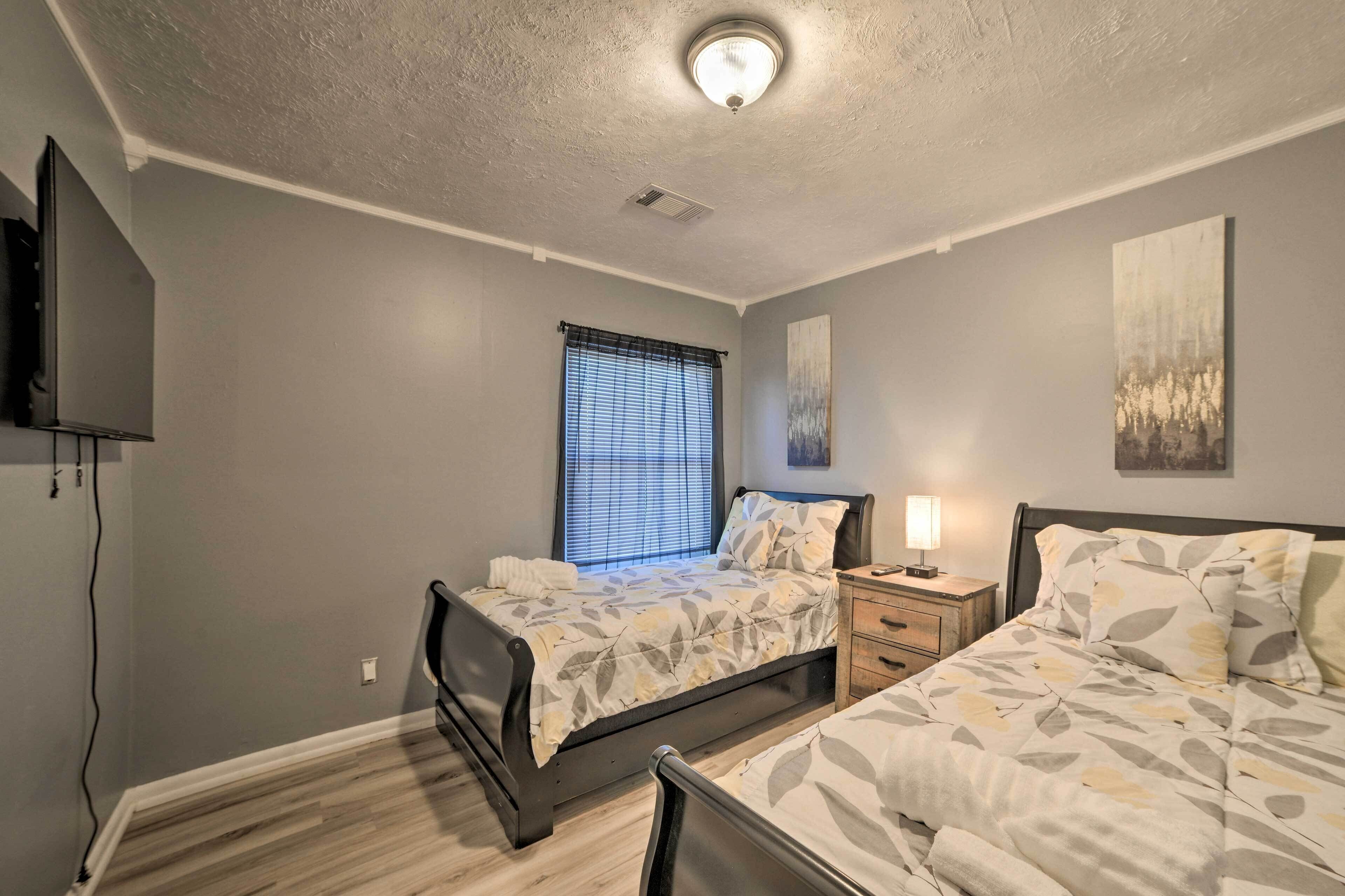 A bedroom with two beds, a TV mounted on the wall, a window with blinds, and a ceiling light.