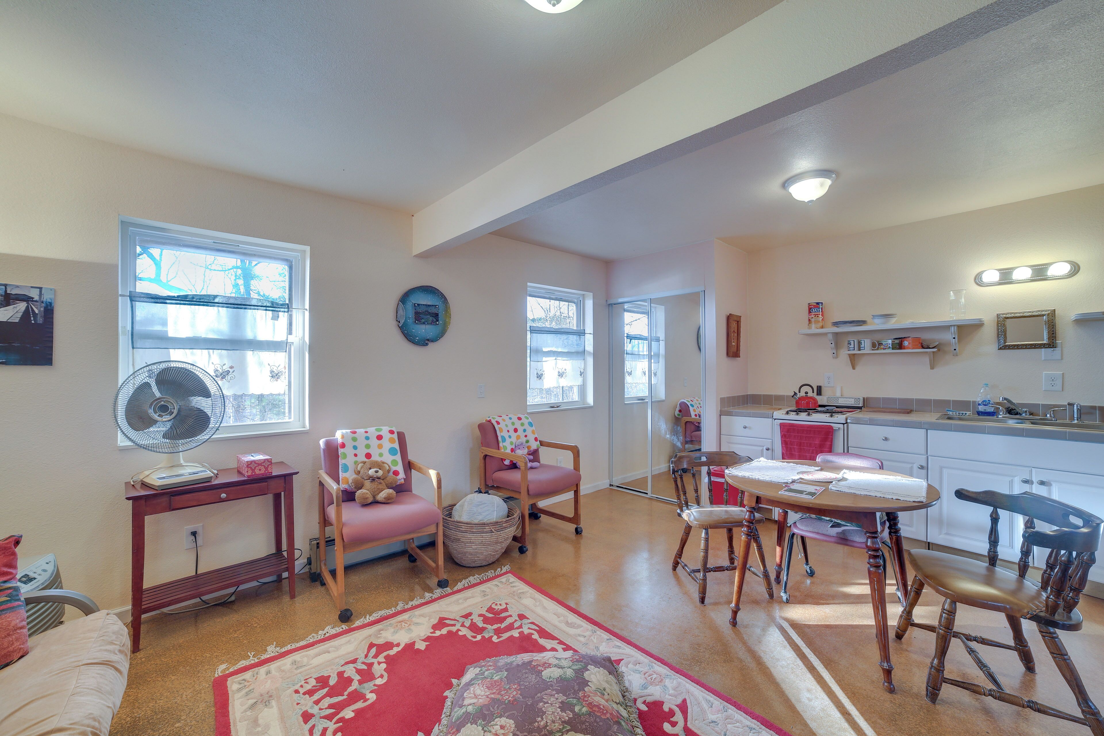 A spacious living room with a large window, a fan, a red rug, and a dining area with wooden chairs and a table.