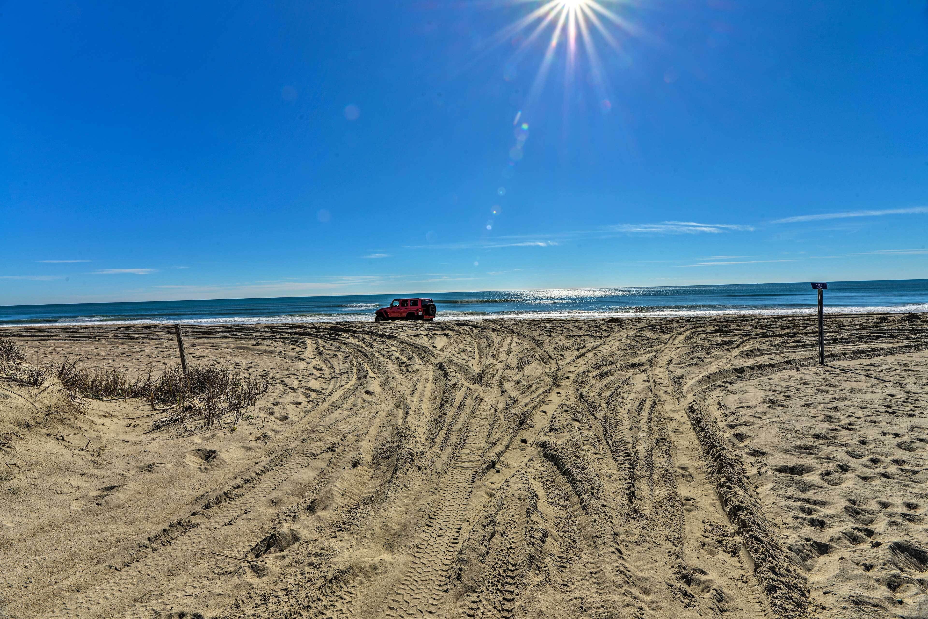 Une plage où l’on voit des traces de pneus menant à un véhicule rouge garé près de l’eau.