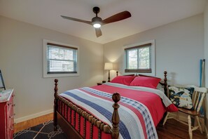 Quarto com cama de madeira, ventoinha no teto, duas janelas com persianas, mesa de cabeceira com abajur e cadeira com almofada decorativa.