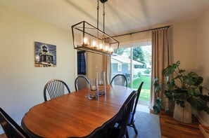 A dining room with a wooden table, chairs, a chandelier, a painting, and a view of the outdoors. - Walk to Beach: Lake Michigan Getaway! (Muskegon)