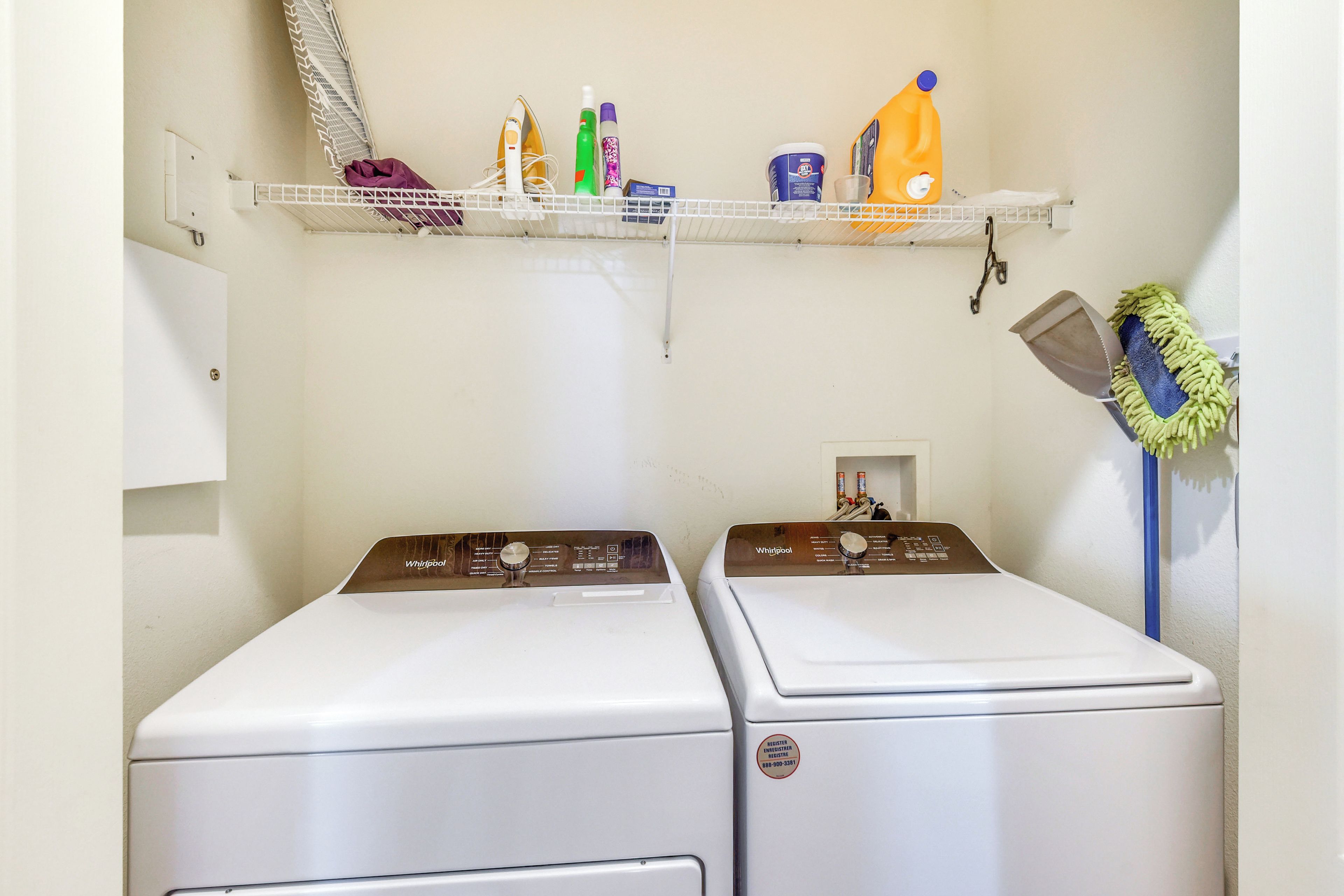 A laundry room with two front-loading washers, a shelf above containing cleaning supplies, and a mop on the right.