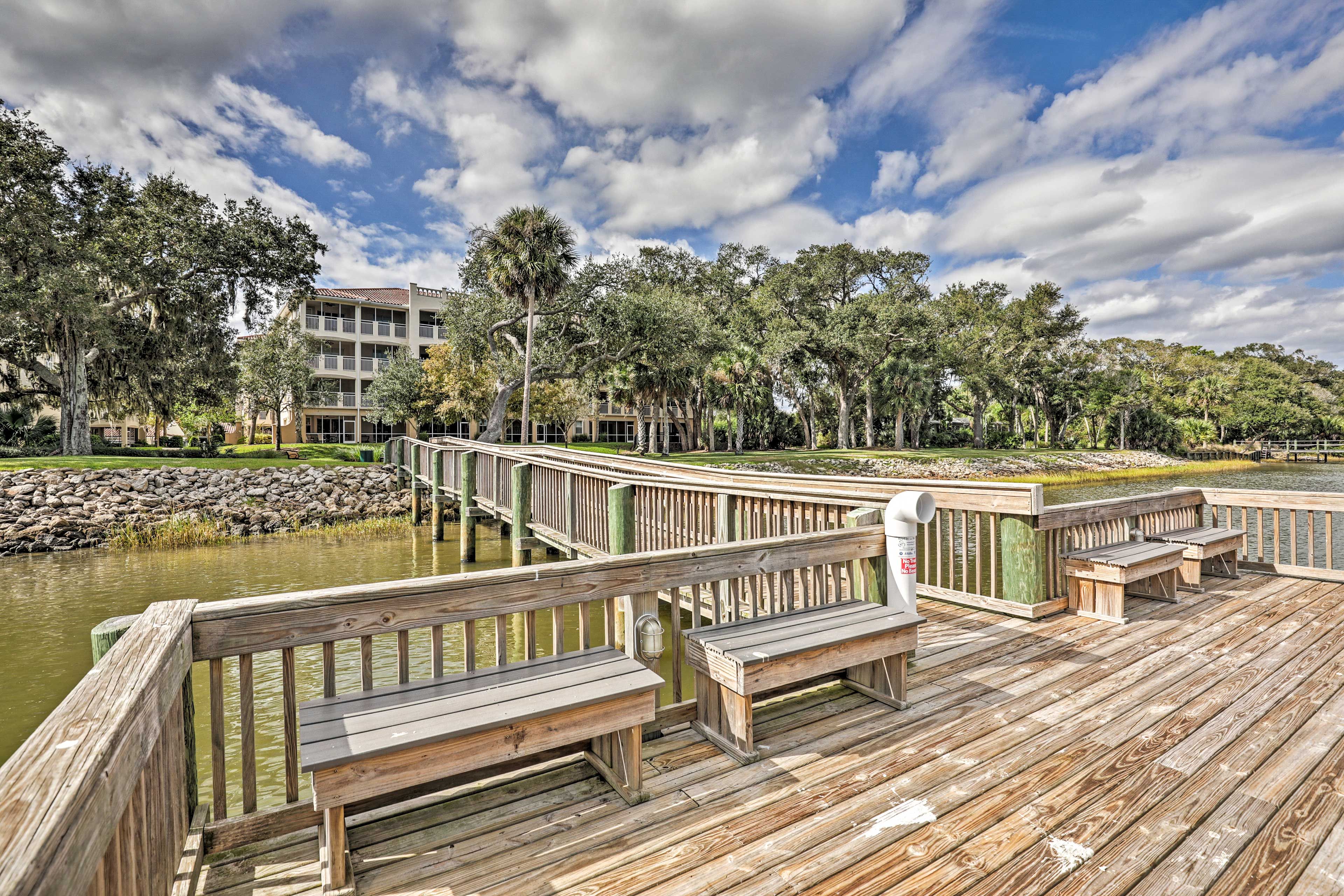 A wooden deck with benches overlooking a body of water, surrounded by trees and a building in the background.