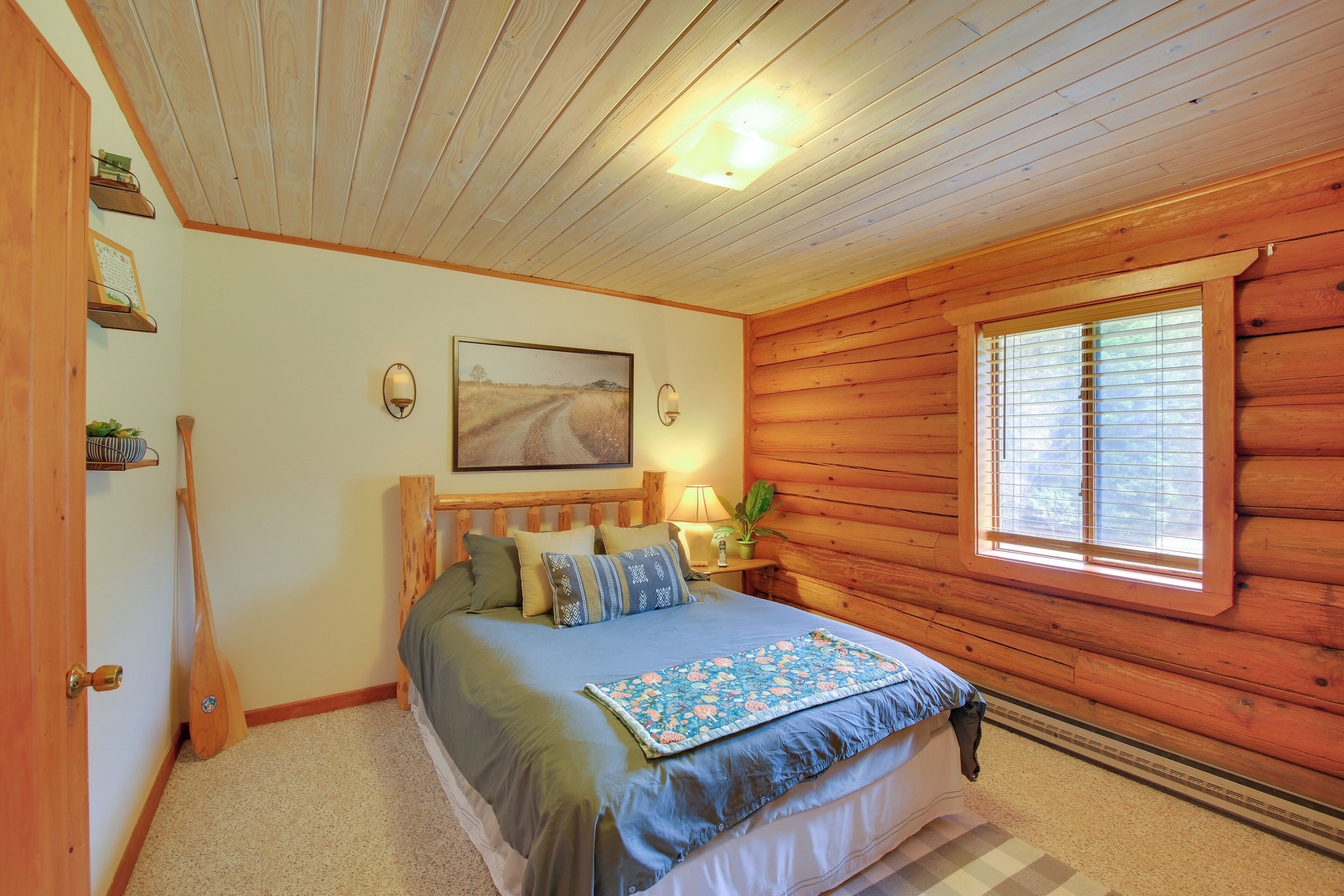 A cozy log cabin bedroom with a bed, pillows, a nightstand with a lamp, a window with blinds, and a painting on the wall.
