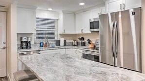 A modern kitchen with white cabinets, a stainless steel refrigerator, a marble countertop, and a window with blinds. - Oceanfront Retreat w/ Beach & Resort Access! (Daytona Beach Shores)