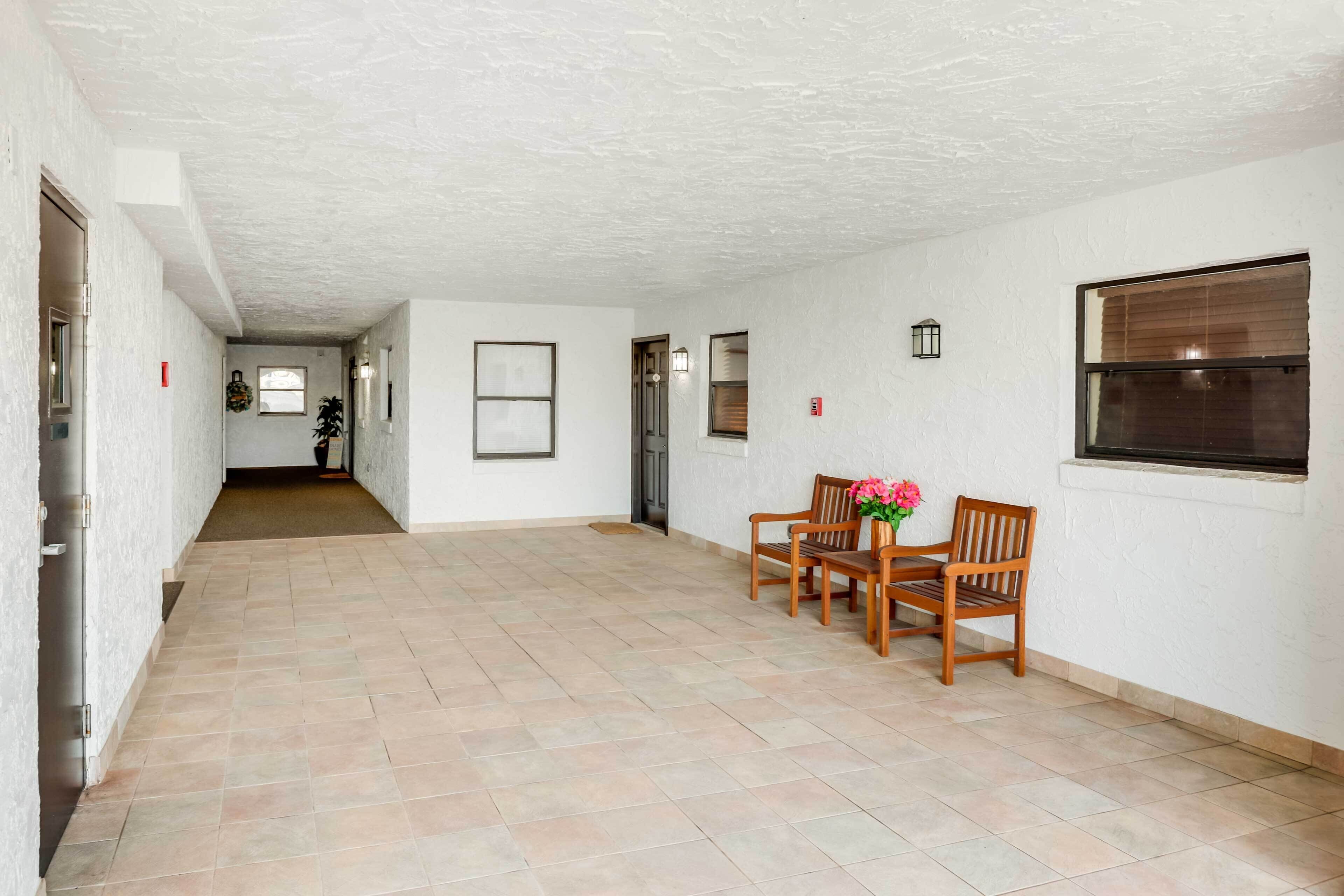 A spacious hallway with a tiled floor, wooden chairs, a small table with a vase of flowers, and a window with blinds.