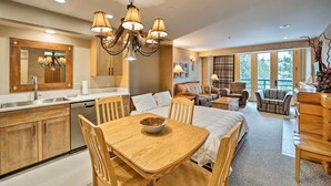A spacious living area with a large window, a dining table with chairs, a sofa, and a kitchen area with wooden cabinets and a chandelier. - Big Sky Condo w/ Mountain Views, Hike, Ski & Fish! (Big Sky)