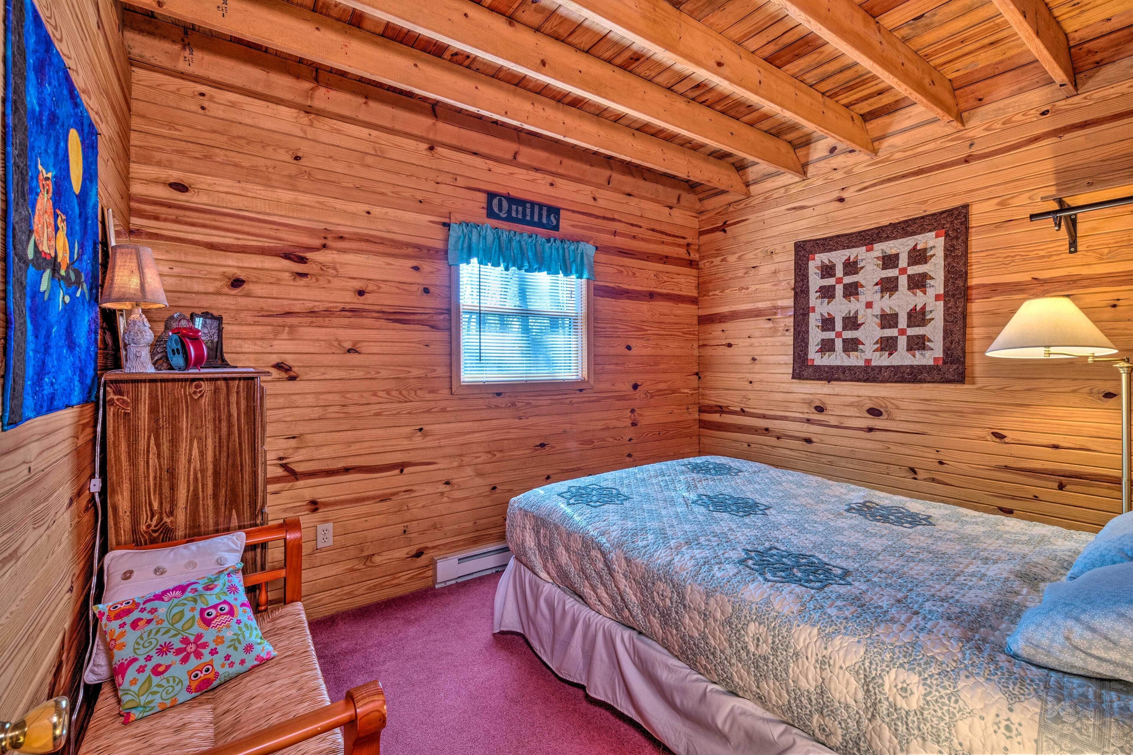A cozy room with wooden walls, a bed with a quilt, a window with blue curtains, a quilted wall hanging, and a lamp.