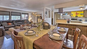 A dining area with a wooden table set for six, surrounded by wooden chairs. A kitchen with a yellow backsplash and modern appliances is visible in the background. - Rustic Western Home: Walk to Winchester Lake! (Winchester)