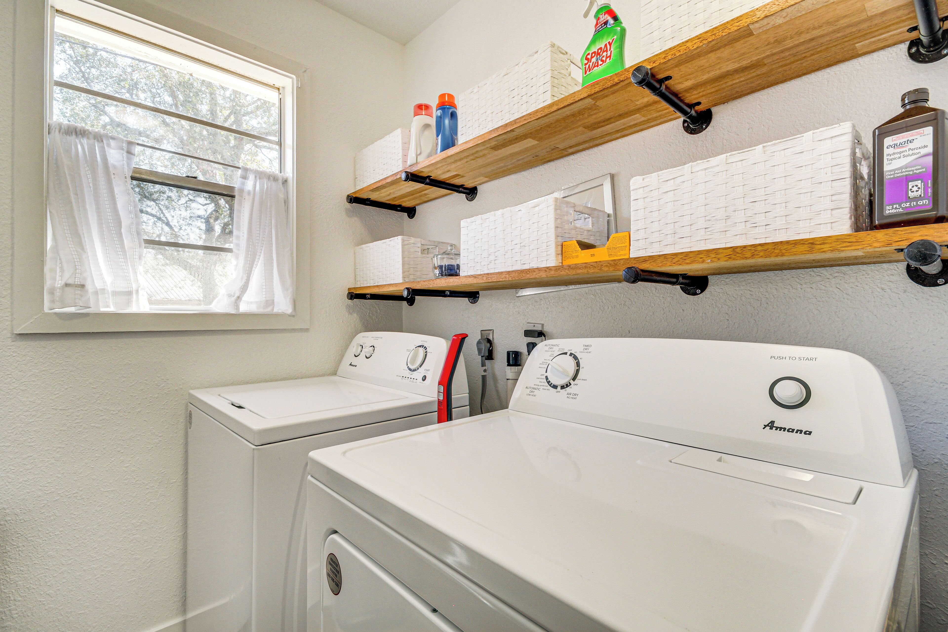 A laundry area with two white Amana washing machines, a window with white curtains, and wooden shelves above holding various items including a spray bottle and a bottle of Equate detergent.