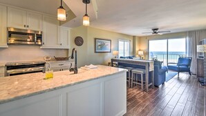 A kitchen with white cabinets, a stainless steel microwave, and a hanging light fixture. There is a dining area with a table and chairs, and a view of the ocean through large windows. - Luxe Oceanfront Condo w/ Pool, Beach Access + Gear (Ormond Beach)