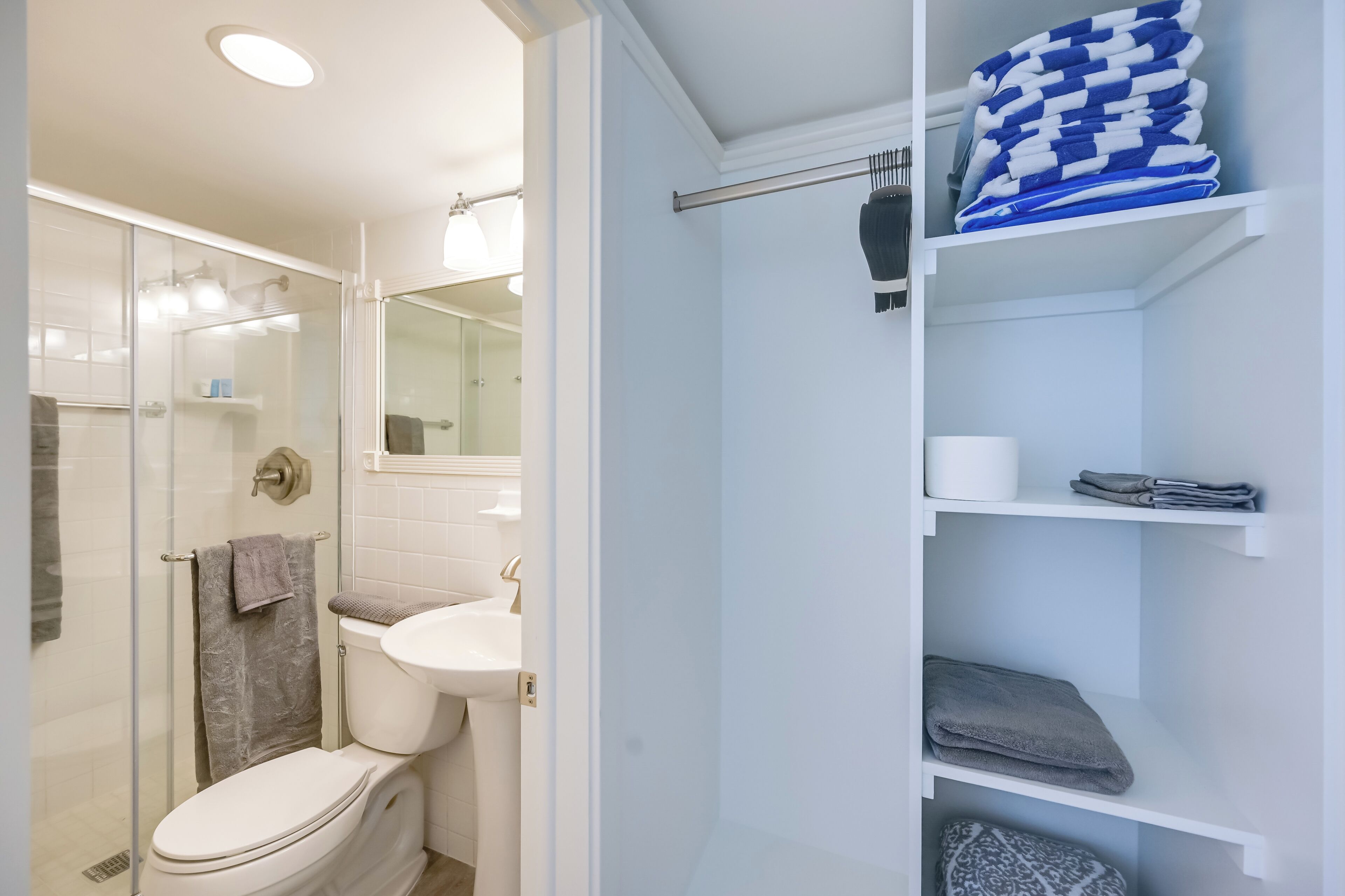 Et moderne badeværelse med glasbrusekabine, hvid vask og toilet. Der er hylder med sammenfoldede håndklæder og en håndklædestige.