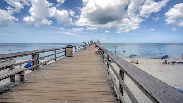 Eine Holzstege führt zu einem Strand mit Sonnenschirmen und Menschen.