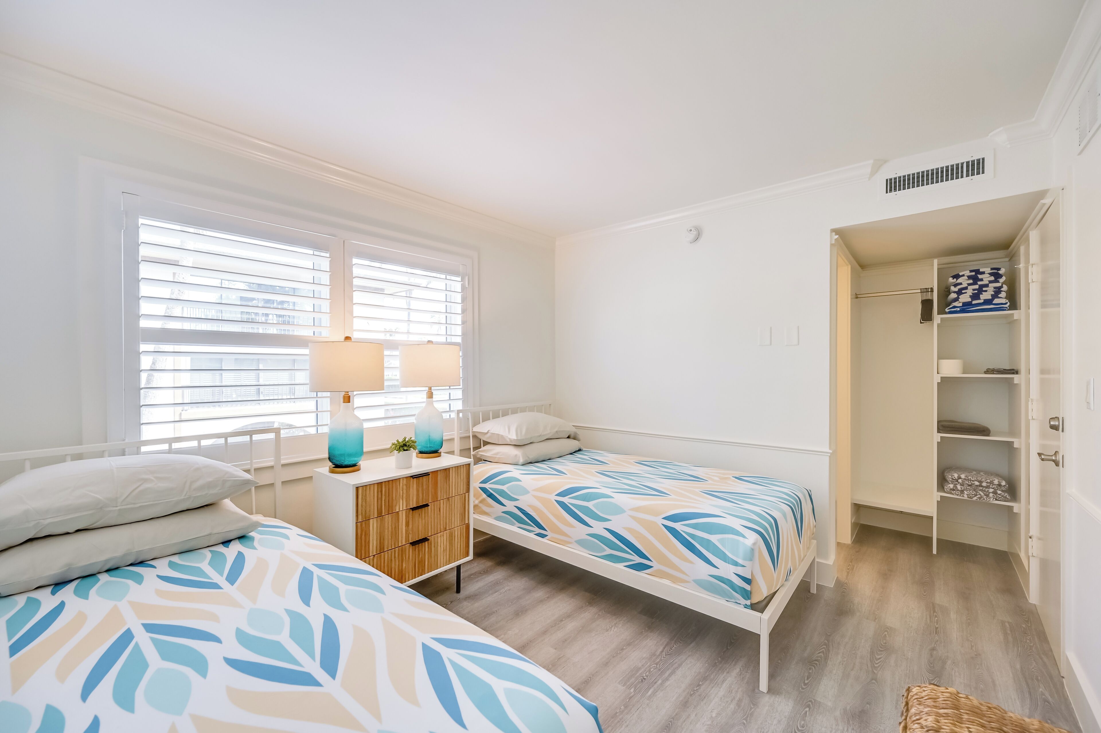 Une chambre avec deux lits, une table de chevet avec une lampe, une armoire et une fenêtre avec des stores.