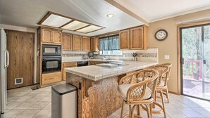 A kitchen with wooden cabinets, a central island with bar stools, a built-in oven, and a large window. - Deck & Hot Tub: Rustic Groveland Home! (Groveland)
