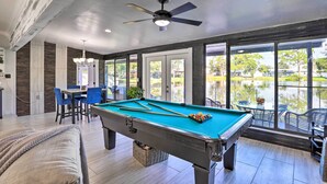 Une table de billard avec des queues et des billes, un ventilateur de plafond et une vue sur un lac par de grandes fenêtres.