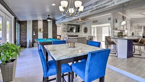 Une cuisine moderne avec un îlot central, un coin repas avec des chaises recouvertes de tissu bleu et, au fond, une table de billard.