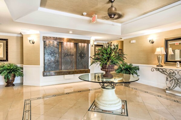A lobby with a glass-top table, potted plants, a wall-mounted light fixture, and an exit sign.