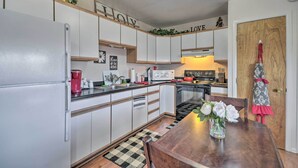 A modern kitchen with white cabinets, a refrigerator, a microwave, and a stove. There is a wooden table with a vase of flowers and a red apron hanging on the door. - 'cedar Mountain Hideout' By Cody Night Rodeo! (Cody)