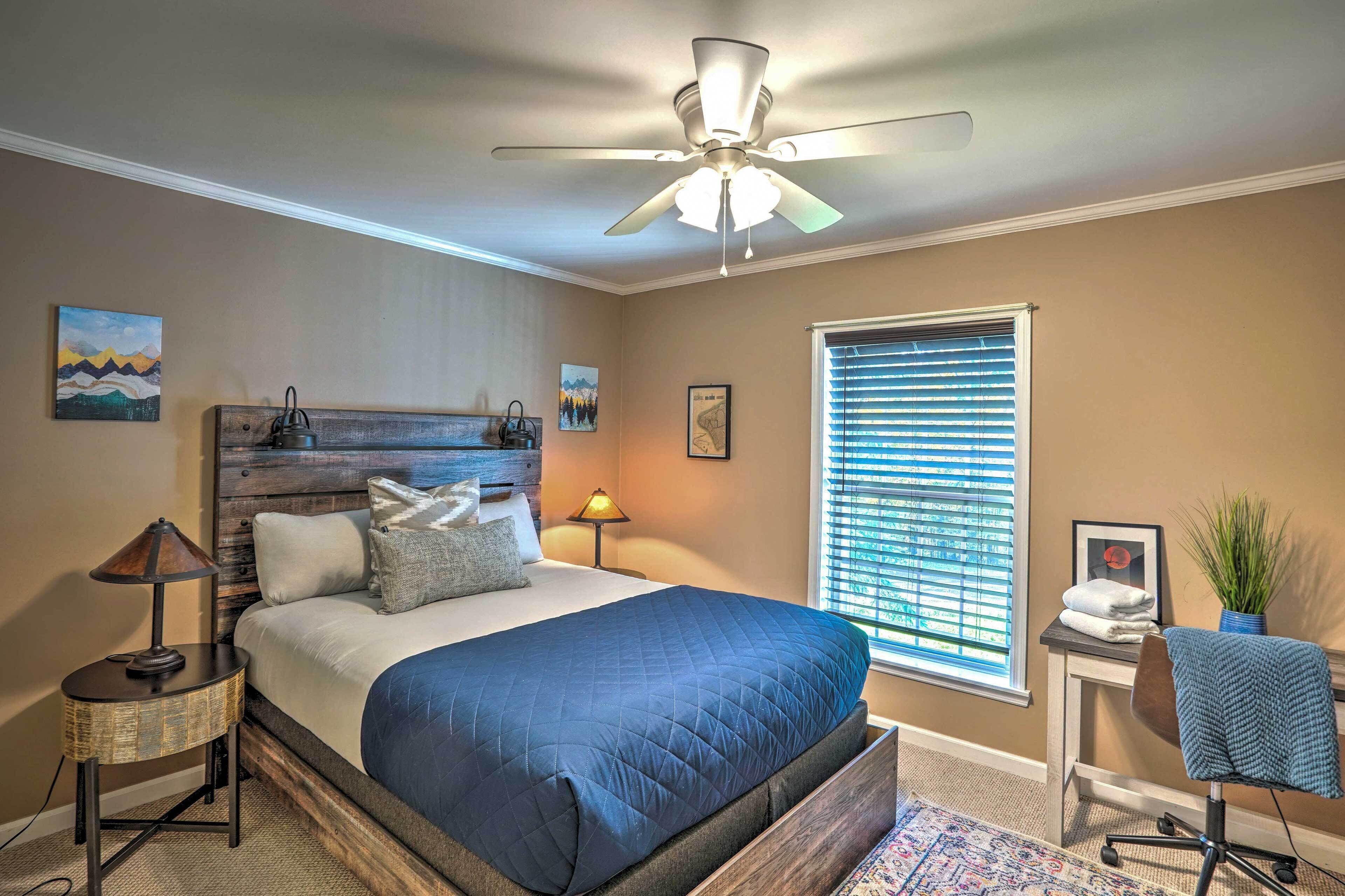 A bedroom with a bed, a ceiling fan, a window with blinds, a nightstand, a lamp, a chair, and a small table with a plant.
