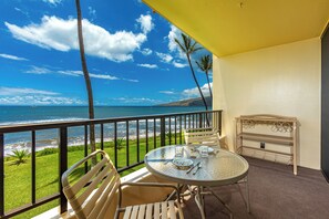 Un balcon avec une table et des chaises en verre, donnant sur une plage bordée de palmiers et sur l’océan.