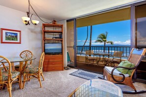 A balcony with a view of the ocean, wicker furniture, a glass table, and a bookshelf. - Beachfront Paradise with A/C - Sugar Beach Resort #227 - Stunning Ocean Views! (Kihei, Maui)