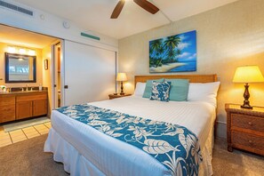 Une chambre d’hôtel avec un grand lit, une commode en bois, une table de chevet avec une lampe et un tableau représentant une scène de plage au mur.