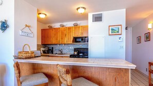 A kitchen with wooden cabinets, a white countertop, and two wicker chairs. - Beachfront Paradise with A/C - Sugar Beach Resort #227 - Stunning Ocean Views! (Kihei, Maui)