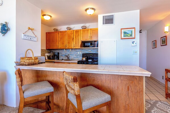 A kitchen with wooden cabinets, a white countertop, and two wicker chairs. - Beachfront Paradise with A/C - Sugar Beach Resort #227 - Stunning Ocean Views! (Kihei, Maui)