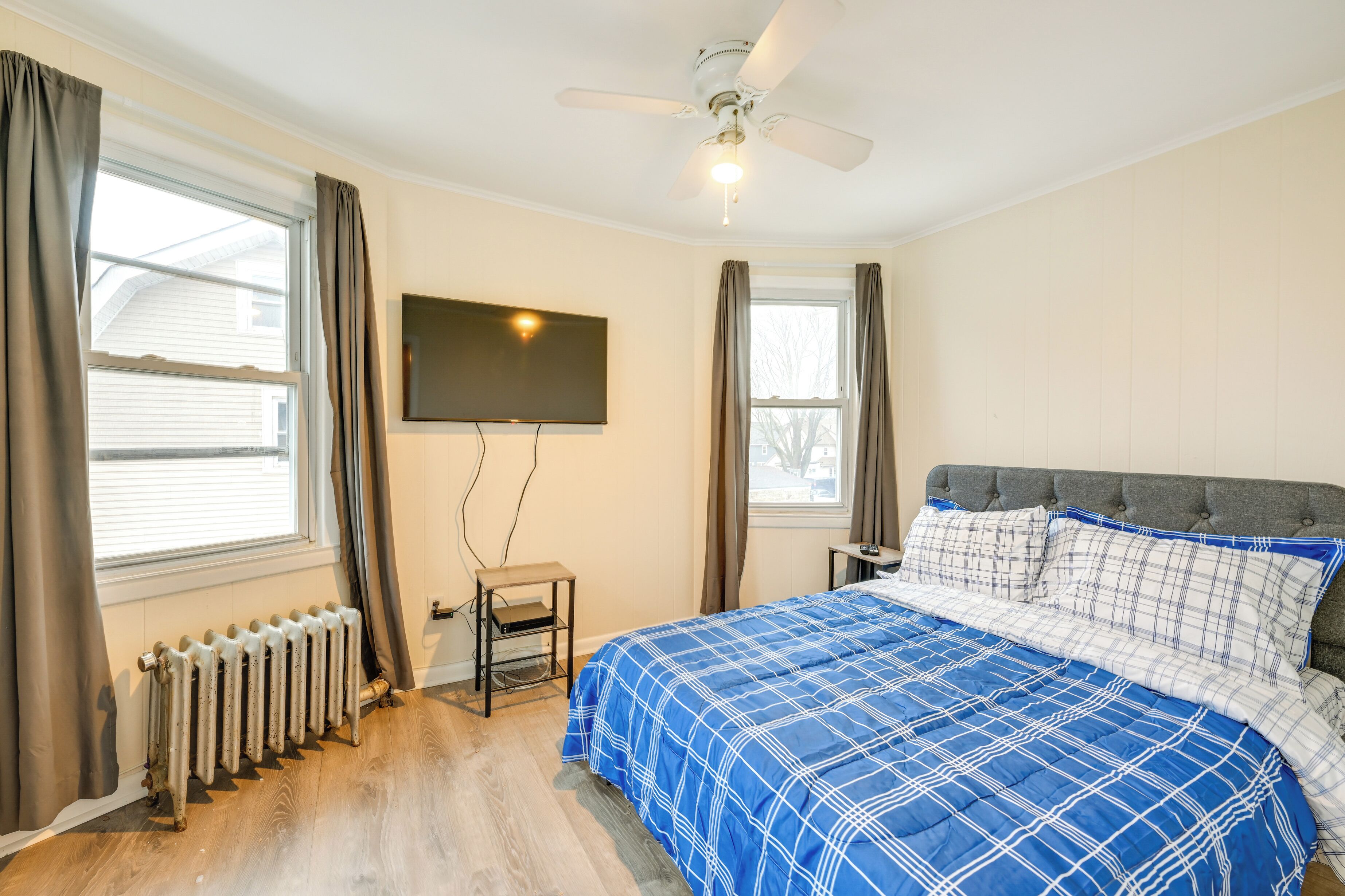 Une chambre à coucher avec un lit, un ventilateur de plafond, un téléviseur fixé au mur et un radiateur.