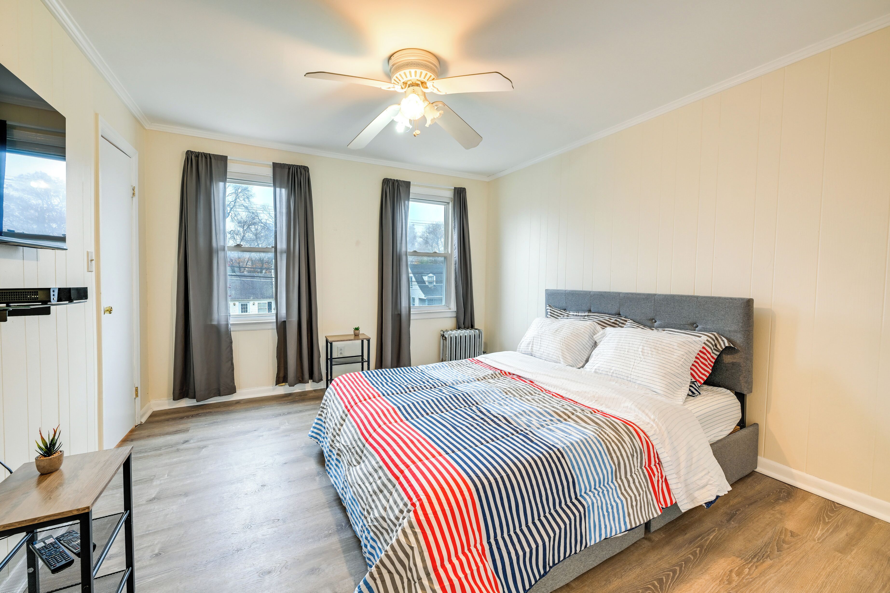 Ein Schlafzimmer mit Bett, Deckenventilator, Fenster mit Vorhängen und einem kleinen Tisch mit Pflanze.