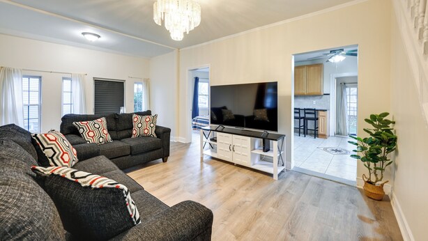 Een moderne woonkamer met een grote flatscreen televisie, grijze banken met gekleurde kussens, een houten salontafel en een kroonluchter.