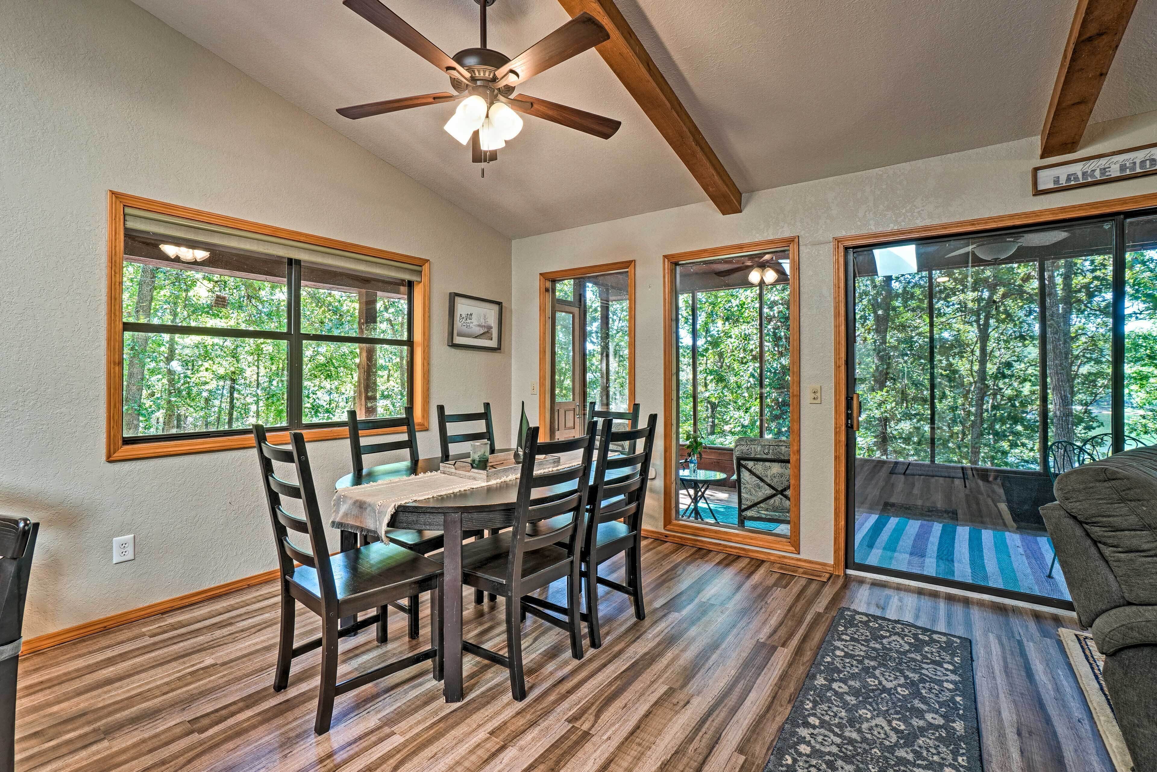 A dining area with a wooden table and chairs, large windows with a view of trees, and a ceiling fan.