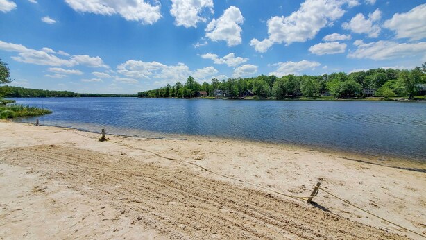 Une plage de sable bordée d’un lac paisible, entourée d’arbres et de maisons qui se dessinent à l’horizon.
