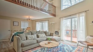 Un salon spacieux avec un plafond haut, une balustrade en bois, une grande fenêtre avec des stores, un tapis à motifs et un coin salon confortable avec un canapé et des fauteuils.