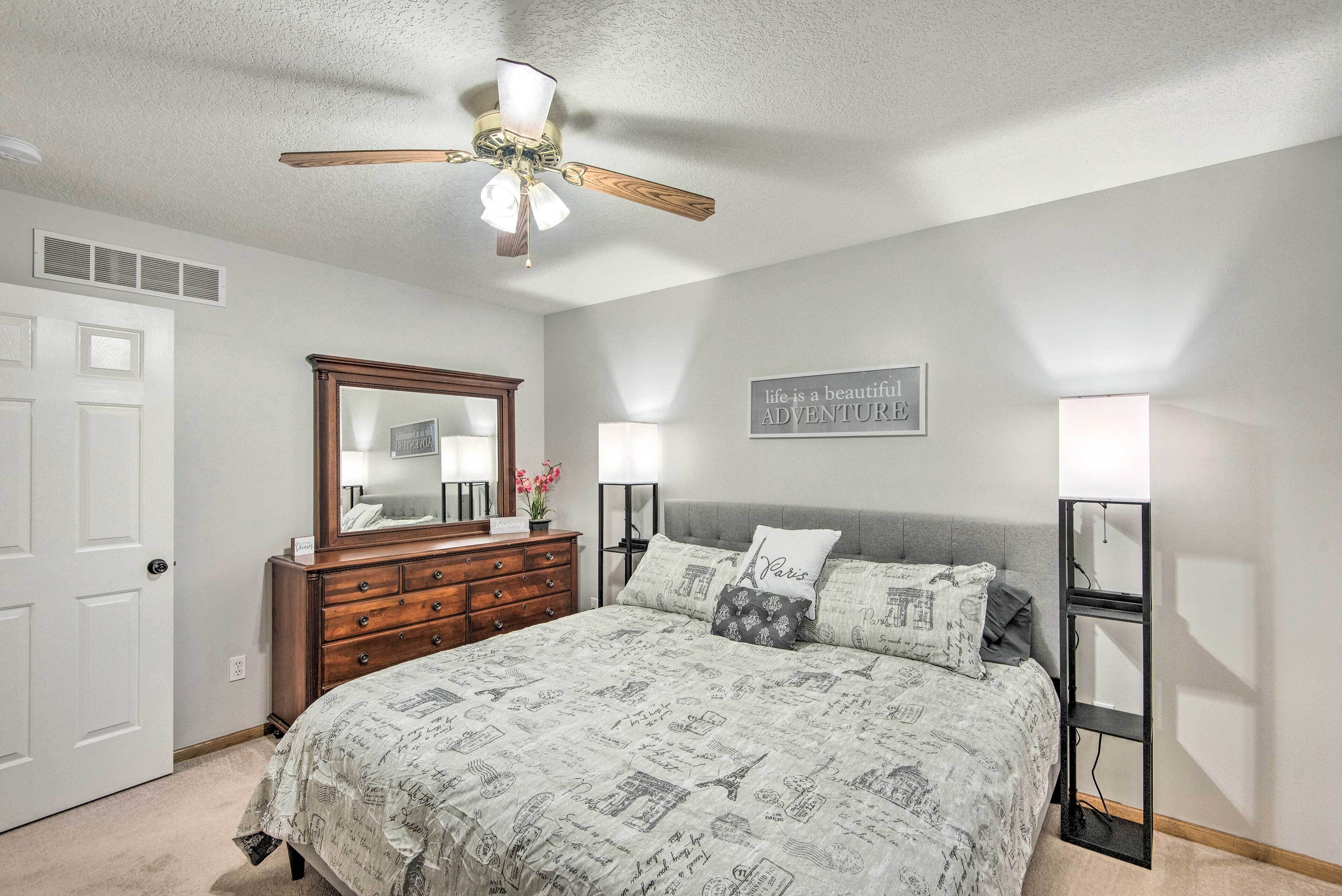 A bedroom with a bed, dresser, mirror, ceiling fan, bedside lamps, and a wall sign.