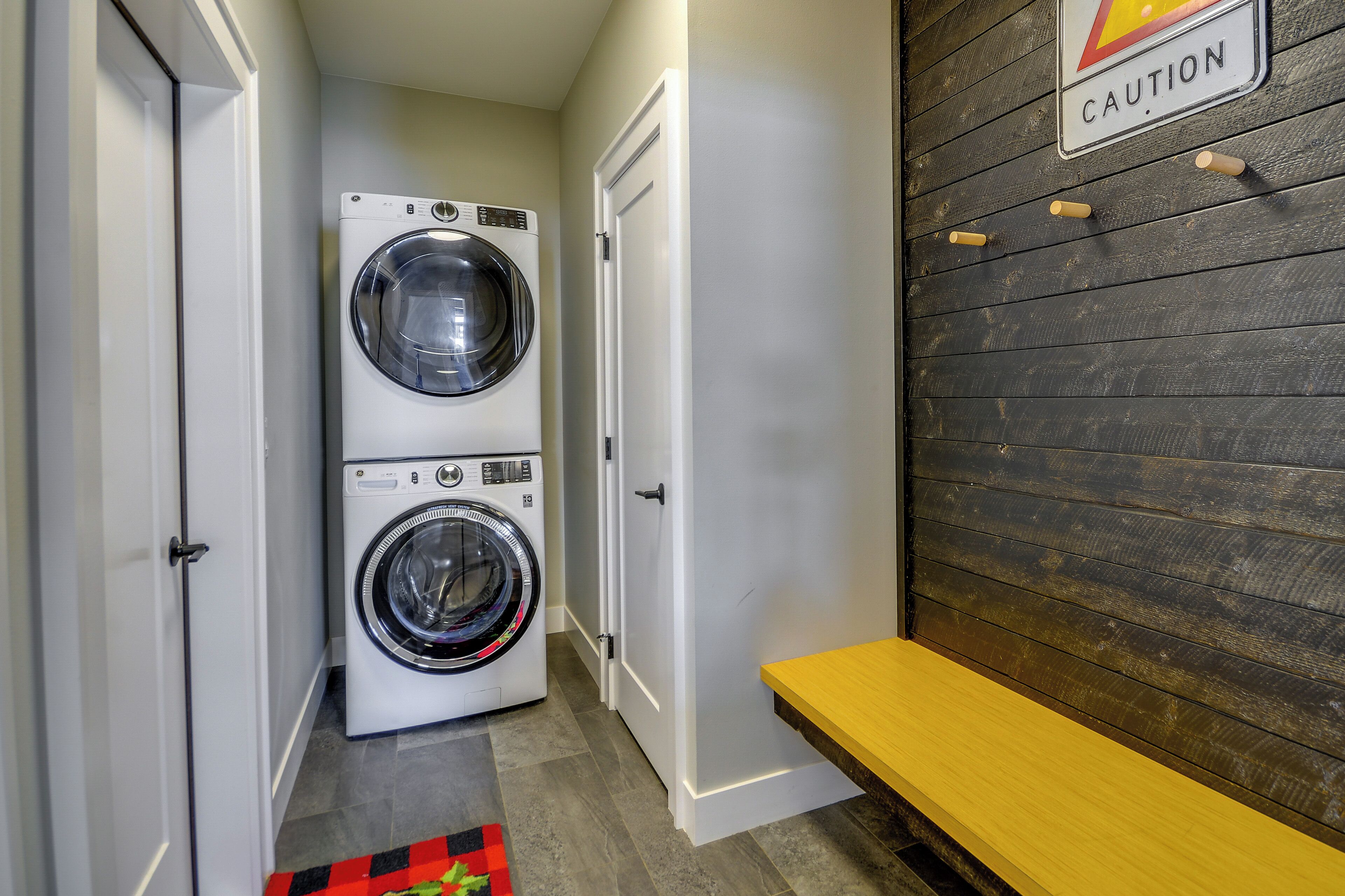A laundry room with two stacked washers and dryers, a caution sign on the wall, and a bench.
