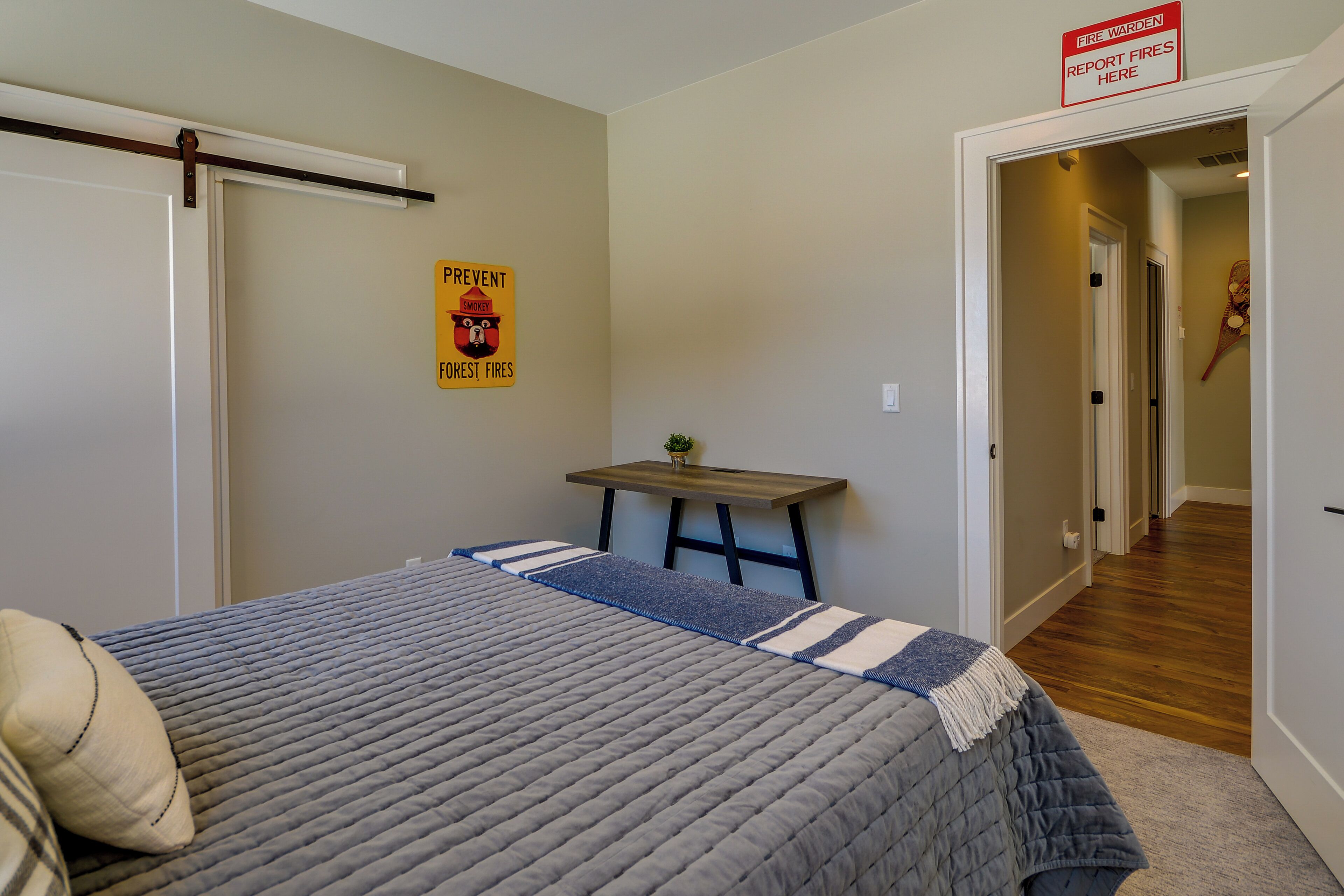 A bedroom with a bed, a small table, a door, and a hallway leading to another room.