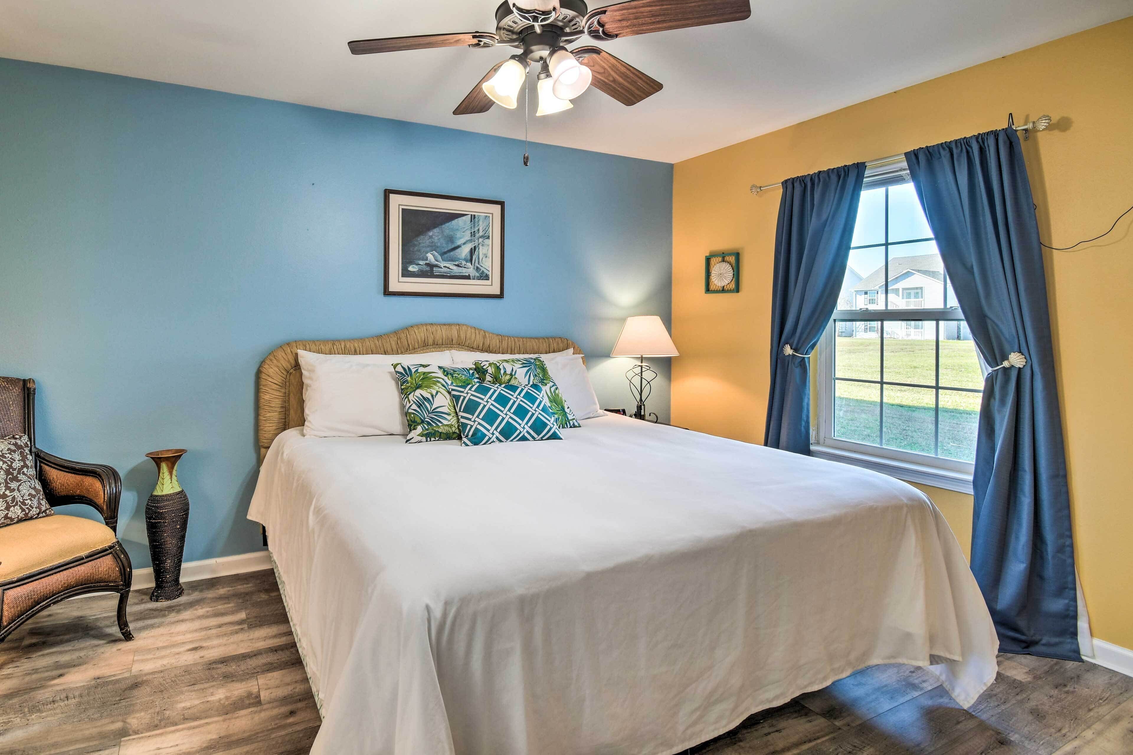 A bedroom with a bed, a ceiling fan, a window with curtains, a lamp, and a framed picture on the wall.