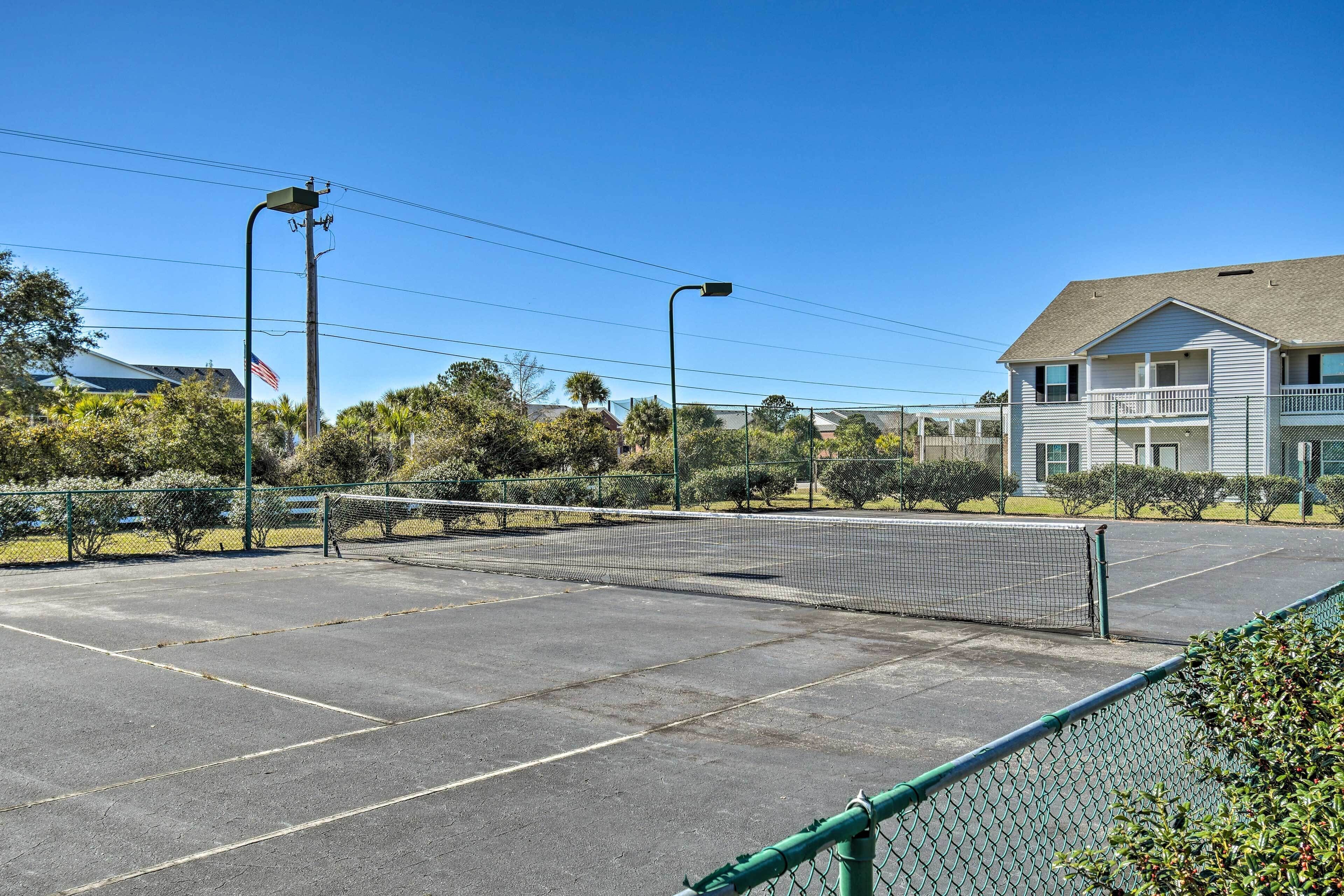 An outdoor tennis court with a net, surrounded by a green fence, and a building with a balcony in the background.
