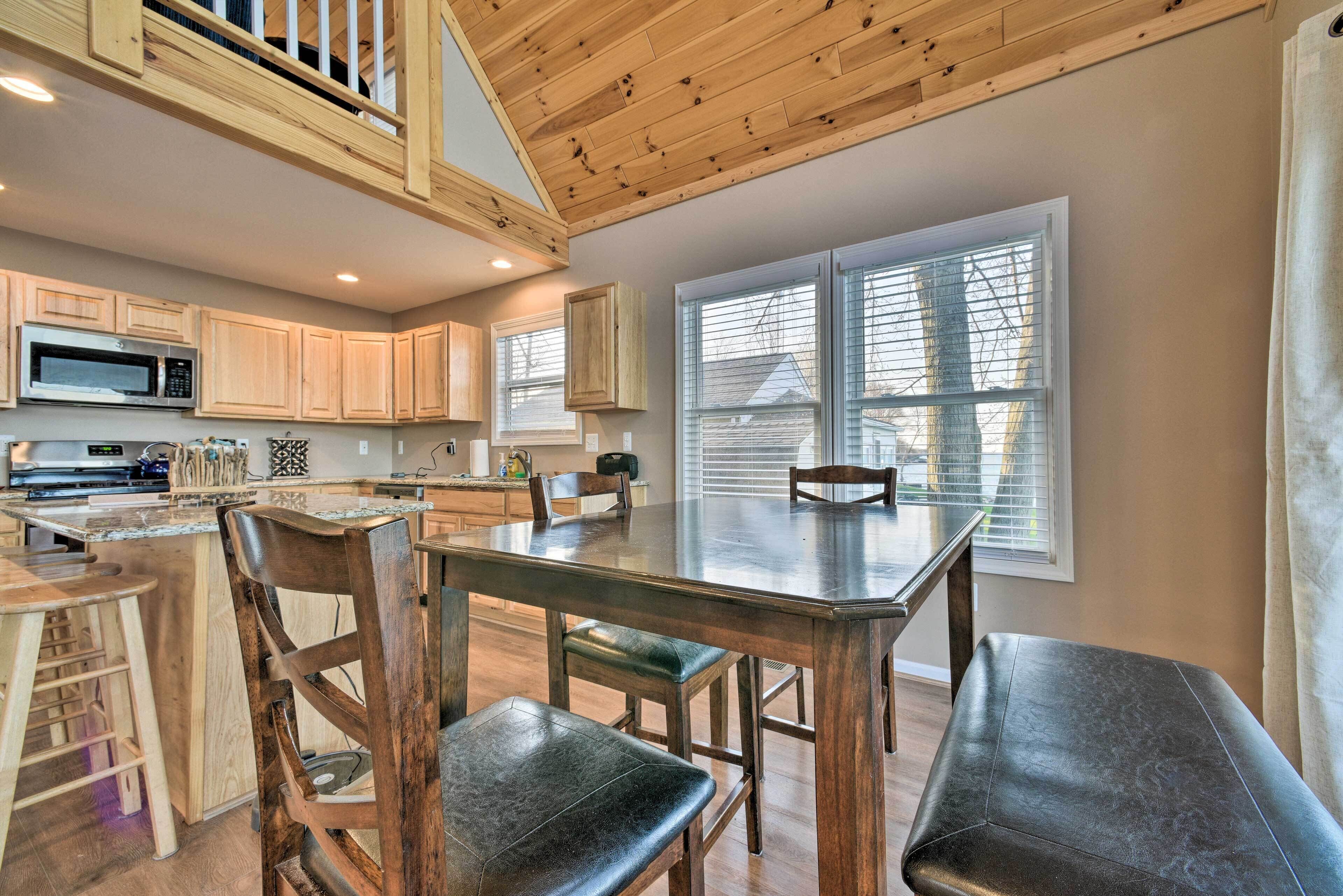 A kitchen with wooden cabinets, a large island with stools, a dining table, and a view of the outside through large windows.