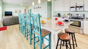 Una cocina moderna con armarios blancos, encimera de granito y electrodomésticos de acero inoxidable. Hay sillas color turquesa alrededor de una isla central con taburetes, y un televisor está montado en la pared.