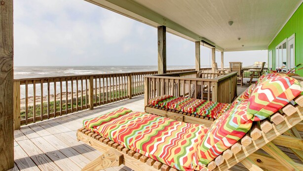 Terraza de madera con dos sillones reclinables acolchados y una mesa, con vista a una playa.