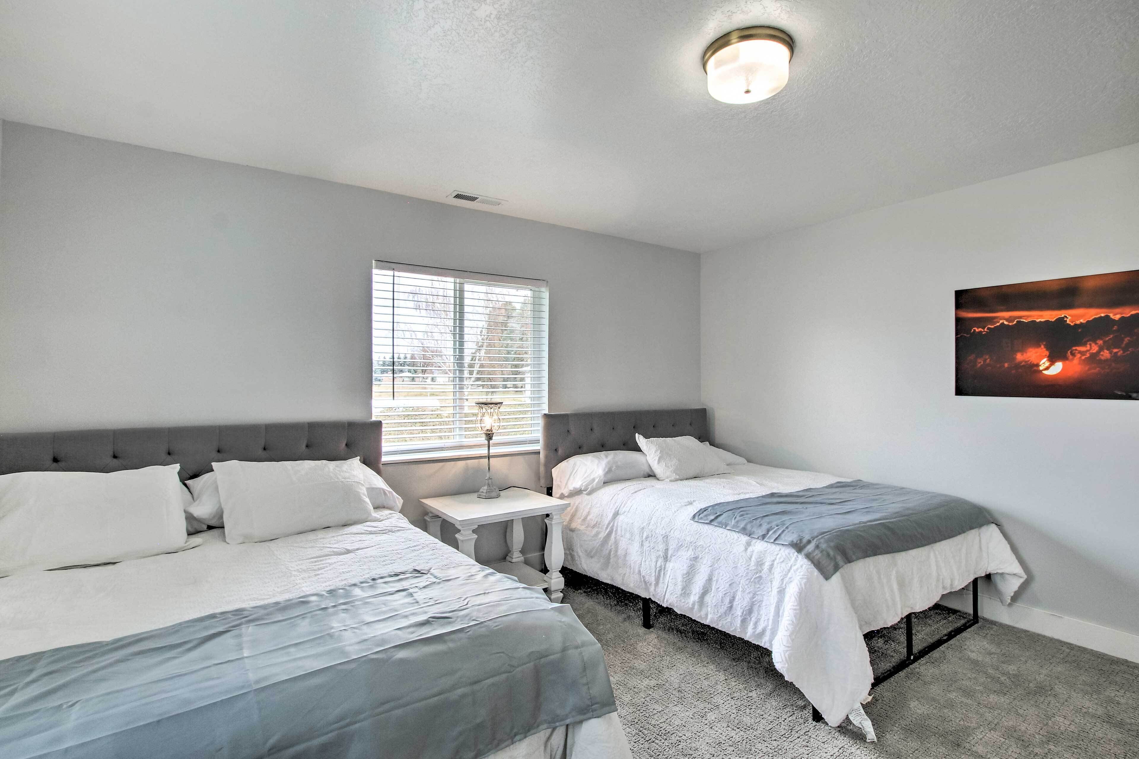 A bedroom with two beds, a window with blinds, a nightstand with a lamp, and a painting on the wall.