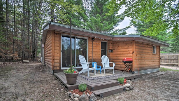 Une cabane en rondins dotée d’une terrasse, de deux fauteuils Adirondack, d’un barbecue et d’un grand bac à fleurs rempli de plantes.