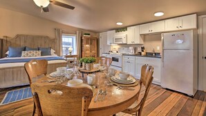 Une chambre confortable avec un lit, une table de chevet et une fenêtre. La pièce comprend une table à manger en bois pour quatre personnes, avec des chaises, un ventilateur de plafond et un coin cuisine avec des placards blancs, un réfrigérateur et un four à micro-ondes.