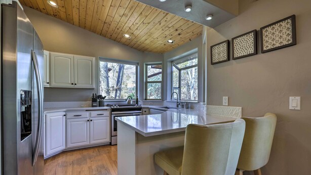 A modern kitchen with white cabinets, a stainless steel refrigerator, a wooden ceiling, and a large island with two chairs. - Cozy Carbondale Escape w/ Deck & Grill! (Carbondale)