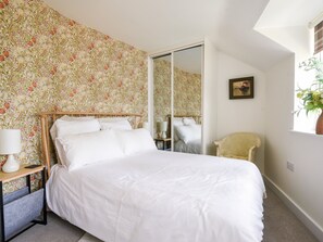 A bedroom with a bed, a nightstand, a chair, a mirror, and a window with a plant. - Conybeare Cottage (Lyme Regis)