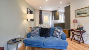 Uma sala de estar com um sofá azul, uma mesinha de centro com abajur, uma cozinha ao fundo e um quadro na parede.